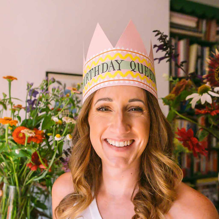 Woman wearing a 'Birthday Queen' party hat with flowers and books in the background
