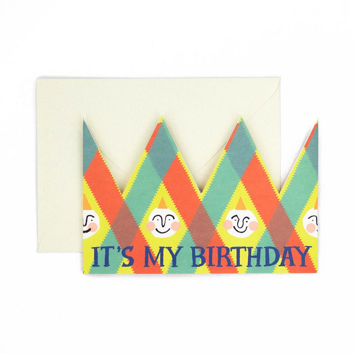 Birthday card with a colorful harlequin pattern and smiling illustrated faces, reading "it's my birthday" placed in front of a cream envelope