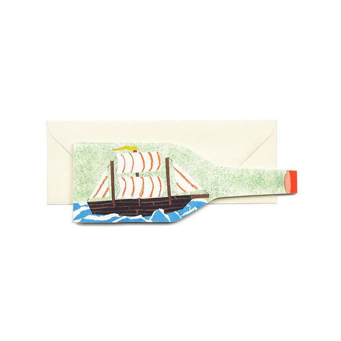 Greeting card shaped like a bottle, featuring an illustration of a ship in a bottle, shown with a long cream envelope.